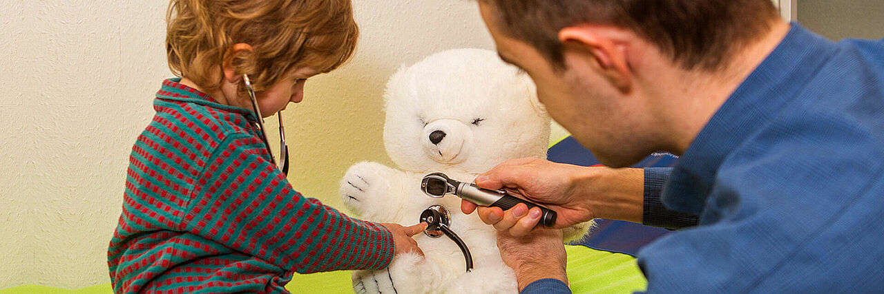 Arzt und kleiner Junge hören mit einem Stethoskop einen weißen Teddy ab.